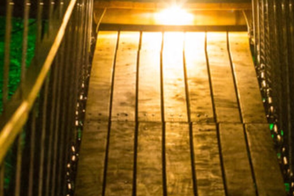 Editorial placeholder image: lantern-lit timber planks of a footbridge, green light at the edge.